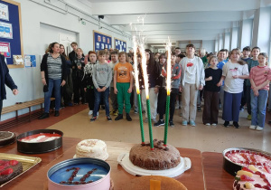 Na zdjęciu widać szkolny korytarz, w którym zgromadziła się duża grupa dzieci wraz z kilkoma dorosłymi. Wszyscy stoją naprzeciw stołu ustawionego na pierwszym planie i patrzą w jego stronę – prawdopodobnie obserwują jakąś uroczystość. Na stole znajduje się kilka ciast i deserów, m.in. torty i wypieki w okrągłych formach. W centrum stoi czekoladowe ciasto z trzema wysokimi, zielonymi świecami-fontannami, które iskrzą się i emitują jasne płomienie, co sugeruje celebrację. Dzieci są ubrane na co dzień, stoją blisko siebie i wyglądają na zainteresowane oraz podekscytowane. W tle widać ściany z tablicami informacyjnymi i ławkę pod ścianą.