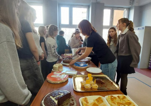 Na zdjęciu widać jasną szkolną korytarz, w którym ustawiono długi stół zastawiony różnymi ciastami i deserami. Przy stole stoi grupa uczniów – głównie nastolatków – którzy ustawili się w kolejce i czekają na swoją porcję. Na pierwszym planie widać różne wypieki: pokrojone kawałki ciasta na tackach, tort czekoladowy z wyciętym fragmentem oraz inne domowe ciasta. Na środku stołu stoi kobieta, która kroi i rozdaje ciasto na talerzyki, obsługując uczniów. Uczniowie stoją blisko siebie, rozmawiają i przyglądają się serwowaniu deserów. W tle znajdują się duże okna wpuszczające dużo światła dziennego, co rozjaśnia całe pomieszczenie.