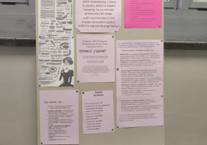 Korytarz szkolny. W tle okna. 1 stojak z tablicą zawierającą ciekawostki o języku polskim.