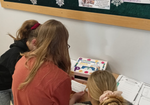 Trzy uczennice są przy stoliku. Jedna z nich siedzi na krześle. Na stoliku znajdują się krzyżówki i rebusy oraz pudełko z rymowankami. Dziewczynki czytają kartkę z wylosowaną rymowanką.