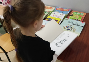 Uczennica ubrana w czarną koszulkę i czarne spodnie w drobne wzorki stoi przy stoliku, na którym leżą książki z wierszykami i rymowankami. Dziewczynka czyta wiersz o bałwanku.