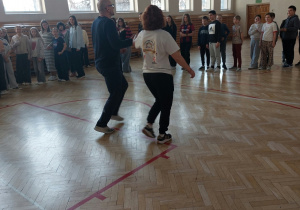 Nauczyciel i nauczycielka stoją na środku sali gimnastycznej. Pokazują uczniom, jak należy tańczyć belgijkę. Dzieci ustawione w parach po okręgu uważnie się przyglądają i zapamiętują po kolei wszystkie ruchy.