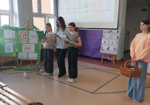 Trzy uczennice stoją na środku i czytają. W tle widać prezentację na temat zdrowia psychicznego. Z boku stoi dziewczynka, która trzyma koszyk z nagrodami.