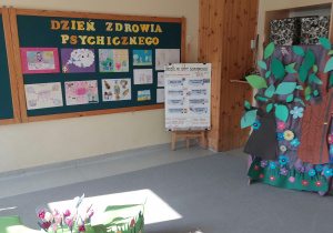 Dekoracja przedstawiająca kwiaty, drzewa. Z tyłu na tablicy wiszą prace konkursowe „Recepta na dobre samopoczucie”. Na górze tablicy widnieje napis Dzień Zdrowia Psychicznego.