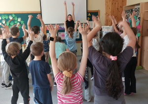 Dzieci tańczą z panią w czarnej bluzce. Mają ręce wysoko podniesione do góry.