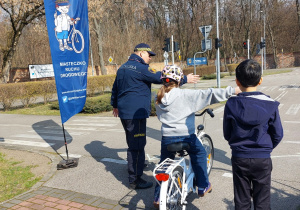Miasteczko Ruchu Drogowego. Tyłem widzimy chłopca w granatowej kurtce, dziewczynkę na rowerze i przedstawiciela Straży Miejskiej, który objaśnia wykonanie zadania.
