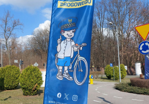 Baner Miasteczko Ruchu Drogowego 986. Na niebieskim tle widnieje rowerzysta.
