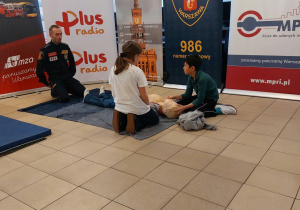 Dzieci – chłopiec i dziewczynka prezentują na fantomie, jak należy udzielić pomocy poszkodowanemu z drgawkami. Obok nich siedzi przedstawiciel Straży Miejskiej i zadaje im dodatkowe pytania.