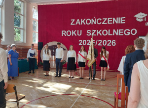 Na środku sali stoi grupa uczniów ubranych w odświętne stroje – białe koszule i ciemne spódnice lub spodnie.
