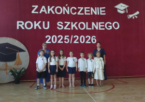 W pierwszym rzędzie stoi siedmioro dzieci ubranych odświętnie – większość ma białe bluzki lub koszule oraz ciemne spódnice lub spodnie. Jedna z dziewczynek ubrana jest w jasną, kremową sukienk. Za dziećmi stoi pani dyrektor i wychowawczyni.