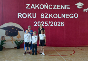 Na pierwszym planie stoi troje uczniów ubranych odświętnie. Dwaj chłopcy mają na sobie białe koszule i ciemne spodnie lub spodenki, a dziewczynka jest ubrana w białą koszulkę polo i ciemną spódnicę. Za uczniami stoi pani dyrektor i wychowawczyni.