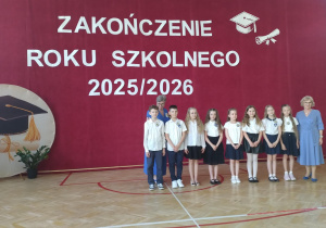 W centralnej części zdjęcia stoi grupa ośmiorga uczniów – dwóch chłopców i sześciu dziewczynek – ubranych odświętnie w białe koszule lub bluzki oraz granatowe lub czarne spodnie i spódniczki. Po prawej stronie grupy znajduje się wychowawczyni ubrana w jasnoniebieską sukienkę. Za uczniami stoi pani dyrektor pochylona nad dwójką dzieci.
