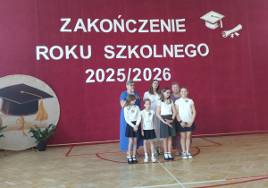 Na środku zdjęcia stoi grupa czterech uczennic. Dziewczynki ubrane są odświętnie – w białe bluzki lub koszulki oraz granatowe lub szare spódniczki. Za uczennicami stoją: pani dyrektor, wicedyrektor i wychowawczyni klasy.