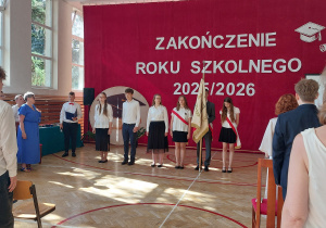 Na środku sali stoi grupa uczniów ubranych w odświętne stroje – białe koszule i ciemne spódnice lub spodnie. Dwoje uczniów ma przewieszone przez ramię biało-czerwone szarfy, a jedna osoba trzyma sztandar szkoły. Po lewej stronie widoczna jest pani dyrektor w niebieskiej sukience oraz uczeń z mikrofonem i teczką, prowadzący uroczystość.