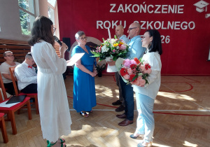 W centrum kadru nauczycielka, trzymając mikrofon i odczytując przygotowany tekst. Przed nią stoją dyrektor szkoły oraz dwie osoby, które wręczają bukiety kwiatów.