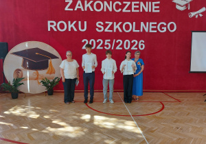 Na zdjęciu widać uroczystość zakończenia roku szkolnego 2025/2026, która odbywa się w sali gimnastycznej. W tle znajduje się duży czerwony baner z napisem „ZAKOŃCZENIE ROKU SZKOLNEGO 2025/2026” oraz dekoracje przedstawiające biret i dyplom. Po lewej stronie ustawiono okrągłą ozdobę z motywem czapki absolwenta i rośliny doniczkowe, a po prawej ekran projekcyjny. Na środku stoi pięć osób. Troje uczniów ubranych w odświętne stroje trzyma w rękach świadectwa. Towarzyszą im dwie nauczycielki – jedna ubrana na biało stoi po lewej stronie grupy, a druga w niebieskiej sukience po prawej.