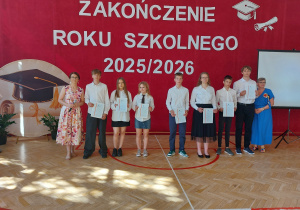 Na środku, na tle czerwonej dekoracji z dużym napisem „ZAKOŃCZENIE ROKU SZKOLNEGO 2025/2026”, stoi grupa uczniów oraz dwie nauczycielki. Uczniowie ubrani są odświętnie – w białe koszule, ciemne spodnie lub spódnice – i trzymają w dłoniach świadectwa. Po lewej stronie grupy stoi nauczycielka w kwiecistej sukience, a po prawej pani dyrektor w niebieskiej sukience.