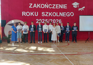 Na środku stoi grupa uczniów wraz z dwiema nauczycielkami. Większość uczniów trzyma w rękach granatowe okładki z napisem „Absolwent”, co sugeruje, że są to absolwenci szkoły. Uczniowie ubrani są odświętnie – część ma białe koszule i ciemne spodnie, inni wybrali bardziej swobodny, ale schludny strój. Po lewej stronie stoi pani dyrektor w niebieskiej sukience, natomiast w centralnej części zdjęcia znajduje się druga nauczycielka ubrana w białą sukienkę. Po prawej stronie ustawiony jest ekran projekcyjny, a po lewej głośnik oraz dekoracja z motywem biretu. Na podłodze widoczne są linie boiska sportowego oraz promienie słońca wpadające przez okna, które rozświetlają parkiet.