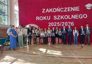 Na środku sali ustawiona jest grupa absolwentów wraz z nauczycielkami. Większość uczniów trzyma granatowe okładki z napisem „Absolwent”, otrzymane podczas ceremonii. Młodzież ubrana jest odświętnie – chłopcy mają białe koszule, marynarki lub eleganckie spodnie, a dziewczęta białe bluzki, spódnice lub eleganckie spodnie. Po lewej stronie zdjęcia stoją dwie kobiety. Jedna z nich ubrana jest w niebieską sukienkę, natomiast druga trzyma mikrofon i odczytuje przygotowany tekst, zwracając się do zgromadzonych uczestników uroczystości. Za nimi widoczni są pozostali uczniowie oczekujący na swoją kolej.