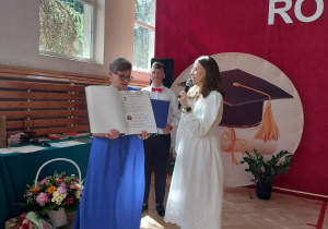 Na pierwszym planie znajdują się dwie nauczycielki. Jedna z nich, ubrana w niebieską sukienkę, prezentuje otwartą kronikę z wpisami i fotografiami uczniów. Obok stoi nauczycielka w białej sukience, trzymająca mikrofon i zwracająca się do uczestników uroczystości. Za nimi widoczny jest uczeń w odświętnym stroju z czerwoną muszką, trzymający niebieską teczkę. Po lewej stronie sali znajdują się duże okna, przez które wpada światło słoneczne, rozjaśniając parkiet i nadając wnętrzu pogodny charakter. Przy stole ustawiono bukiety kwiatów przygotowane z okazji zakończenia roku szkolnego. Całość tworzy uroczystą i podniosłą atmosferę, charakterystyczną dla szkolnych ceremonii podsumowujących rok nauki.