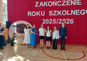 Na tle dużej czerwonej dekoracji z napisem „ZAKOŃCZENIE ROKU SZKOLNEGO 2025/2026” stoi sześć osób ustawionych do pamiątkowego zdjęcia. W centrum znajduje się pani dyrektor w długiej niebieskiej sukni, trzymająca kronikę. Po jej lewej stronie stoi kobieta w białej, eleganckiej sukience i okularach. Po prawej stronie pani dyrektor ustawionych jest troje uczniów w galowych strojach – białych koszulach i ciemnych spódnicach lub spodniach – oraz wysoki uczeń w granatowym garniturze z czerwonym krawatem.