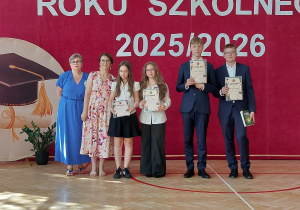 W centrum kadru stoi siedem osób ustawionych do pamiątkowego zdjęcia. Po lewej stronie znajdują się dwie nauczycielki – jedna ubrana w długą niebieską suknię, druga w jasną sukienkę w kwiatowy wzór. Obok nich stoją dwie uczennice w galowych strojach: białych koszulach i ciemnych spódnicach lub spodniach. Obie trzymają w rękach dyplomy. Po prawej stronie widoczni są dwaj uczniowie ubrani w eleganckie granatowe garnitury. Każdy z nich prezentuje otrzymany dyplom, a uczeń stojący skrajnie z prawej trzyma dodatkowo książkę.