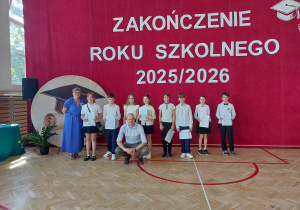 Po lewej stronie stoi pani dyrektor ubrana w długą niebieską suknię. Obok niej ustawionych jest ośmioro uczniów w odświętnych strojach – białych koszulach i ciemnych spódnicach lub spodniach. Część dzieci trzyma w dłoniach świadectwa. Przed grupą, w pierwszym rzędzie, kuca nauczyciel w jasnej koszuli, pozując do wspólnego pamiątkowego zdjęcia.