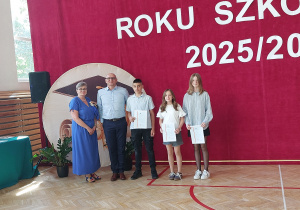 Na pierwszym planie stoi pięć osób. Dwoje dorosłych – pani dyrektor w niebieskiej sukience i pan Sznajder w jasnoniebieskiej koszuli – pozują razem z trojgiem uczniów. Każdy z uczniów trzyma w ręku świadectwo.
