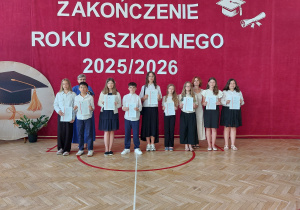Na pierwszym planie stoi ośmioro uczniów ubranych w odświętne stroje. Każdy z nich trzyma w rękach świadectwo. Za uczniami stoją dwie nauczycielki, które wspólnie pozują do pamiątkowego zdjęcia.