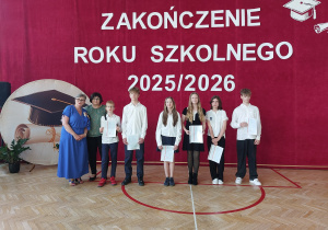 Na pierwszym planie stoi sześcioro uczniów ubranych w odświętne stroje. Większość z nich trzyma w rękach świadectwa, które otrzymali podczas ceremonii. Za uczniami znajdują się dwie nauczycielki, wspólnie pozujące do pamiątkowego zdjęcia.