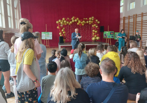 Na zdjęciu przedstawiono uroczystość odbywającą się w szkolnej sali gimnastycznej. W centralnej części fotografii na niewielkiej scenie stoi mężczyzna ubrany w niebieską marynarkę i jasną koszulę. Trzyma w jednej ręce mikrofon, a w drugiej kartkę, z której odczytuje przemówienie, zwracając się do zgromadzonych uczestników wydarzenia. Scena została efektownie udekorowana bordowo-złotą girlandą z balonów oraz kurtyną z ciepłych lampek, tworzącą eleganckie tło. Po obu stronach sceny ustawiono głośniki nagłośnieniowe. W tle widoczna jest bordowa kotara, a po prawej stronie fragment szkolnej dekoracji. Na pierwszym planie znajdują się uczestnicy uroczystości siedzący tyłem do obiektywu.