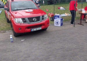 Na pierwszym planie stoi czerwony samochód terenowy z oznaczeniami jednostki ratowniczej oraz sygnałem świetlnym na dachu. Po prawej stronie znajduje się biały namiot, pod którym przygotowano stanowisko informacyjne. Przy stoliku pracują osoby w czerwonych koszulkach, a obok ustawiono pudełka z wyposażeniem medycznym oraz sprzętem do udzielania pierwszej pomocy.