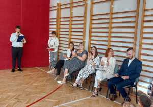 Na krzesłach siedzi grupa nauczycieli, którzy brawami reagują na trwającą uroczystość. Po lewej stronie znajduje się uczeń prowadzący część oficjalną – trzyma mikrofon oraz kartkę z tekstem. Za nim stoi drugi uczeń z instrumentem perkusyjnym. W centrum kadru widoczni są nauczyciele.