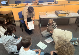 Na zdjęciu widać grupę uczniów uczestniczących w zajęciach edukacyjnych. Dzieci pracują w niewielkich grupach na podłodze, rozwiązując zadania i korzystając z przygotowanych materiałów. Prowadząca nauczycielka prezentuje uczniom kartę z rzymskimi cyframi i ich wartościami.