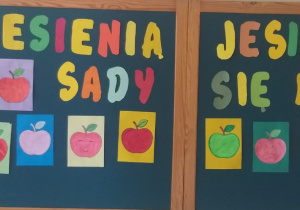Na ciemnozielonym tle widnieje kolorowy napis „Jesienią, jesienią sady się rumienią”. Wokół napisu umieszczone są liczne rysunki czerwonych, zielonych żółtych jabłek przyklejonych do kolorowych prostokątnych kartek. Praca ma wesoły, jesienny charakter i stanowi dekorację świetlicy.