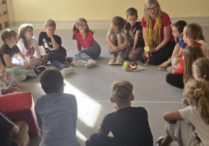 Na zdjęciu widać grupę dzieci siedzących w kręgu na podłodze podczas zajęć świetlicowych. W centrum siedzi nauczycielka prezentująca budowę jabłka i jego gatunki . W tle – na tablicy- znajdują się rysunki dzieci przedstawiające różnokolorowe jabłka.