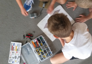 Na zdjęciu widać troje dzieci siedzących na podłodze i wspólnie bawiących się klockami LEGO. Z zainteresowaniem oglądają instrukcję i próbują dopasować poszczególne elementy do budowanej konstrukcji. Są skupione na zadaniu, współpracują ze sobą.