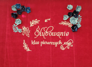 Duża, czerwona dekoracja z napisem „Ślubowanie klas pierwszych”, ozdobiona białymi motywami roślinnymi, kokardami i papierowymi kwiatami w odcieniach błękitu oraz granatu.