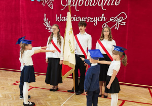 Na zdjęciu widać uroczystość ślubowania uczniów klas pierwszych. Na środku sali gimnastycznej stoi troje starszych uczniów ubranych w białe koszule, czarne spódnice lub spodnie oraz czerwone szarfy przewieszone przez ramię. Pomiędzy nimi znajduje się sztandar szkoły ze złotymi zdobieniami i frędzlami. Przed starszymi uczniami stoją troje młodszych dzieci ubranych w stroje galowe oraz niebieskie birety. Dzieci wyciągają ręce w kierunku uczniów ze szarfami i sztandaru, uczestnicząc w uroczystym elemencie ceremonii. Dziewczynki mają na sobie białe bluzki, granatowe lub czarne spódnice i białe rajstopy, a chłopiec ubrany jest w granatowy sweter i ciemne spodnie. W tle znajduje się duża, czerwona dekoracja z napisem „Ślubowanie klas pierwszych”, ozdobiona białymi motywami roślinnymi, kokardami i papierowymi kwiatami w odcieniach błękitu oraz granatu.