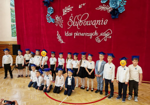 Na zdjęciu widać uczniów klas pierwszych ustawionych w szeregu podczas uroczystości ślubowania. Dzieci ubrane są w białe koszulki lub bluzki, ciemne spódnice i spodnie oraz niebieskie birety. W tle widoczna jest czerwona dekoracja z białym napisem nawiązującym do ślubowania klas pierwszych.